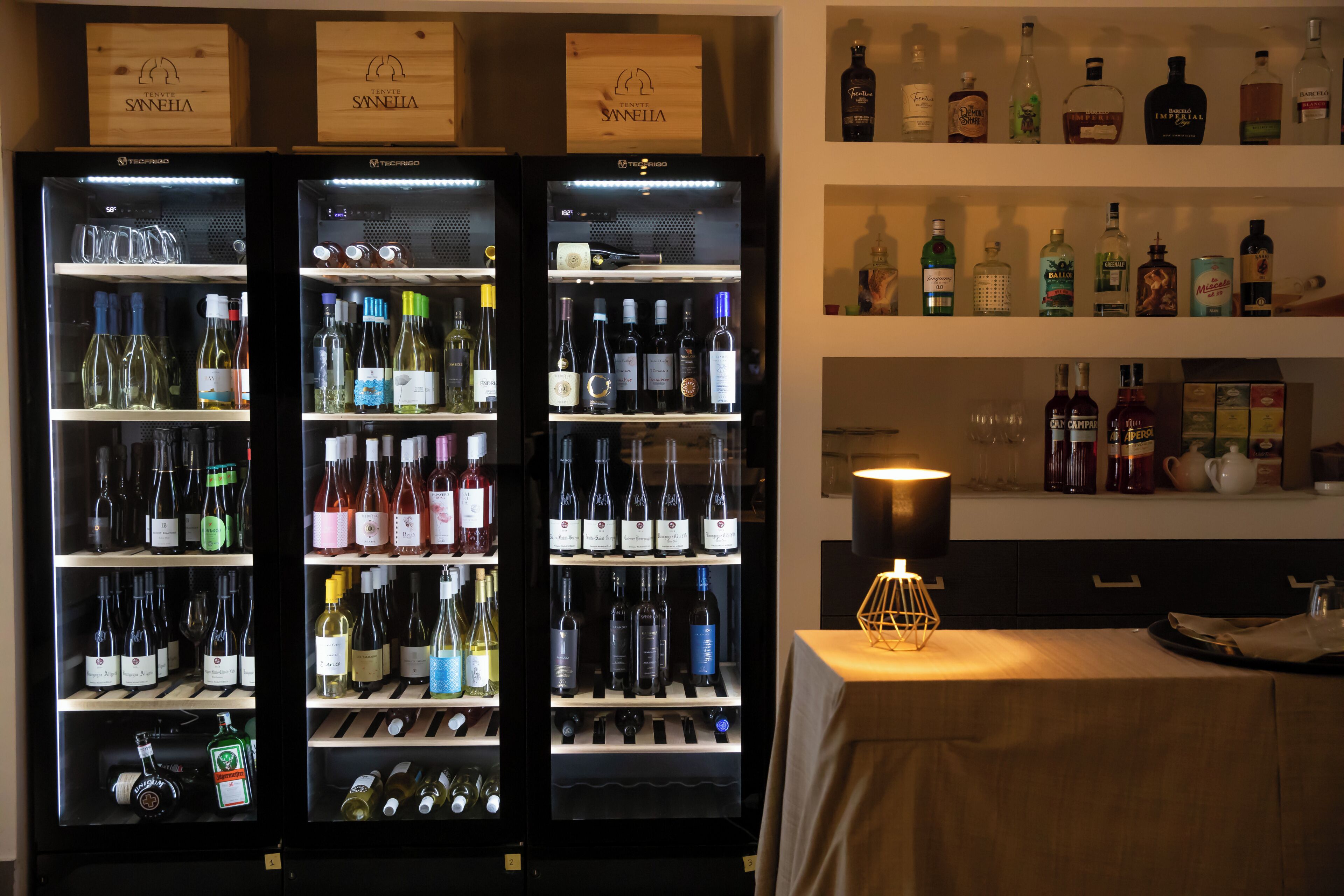Zona de bar con minibar repleto de bebidas alcohólicas, una selección de botellas de vino y una mesa con lámpara.