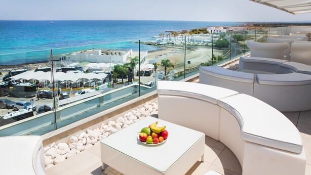 A balcony with a white table and a bowl of fruit, overlooking a beach and a cityscape.