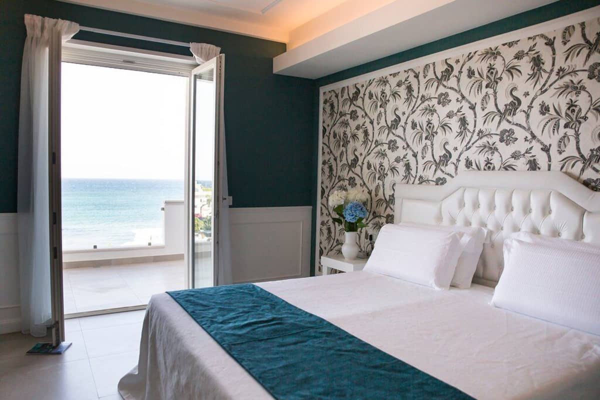 A bedroom with a bed, a nightstand, a window with curtains, and a view of the sea.