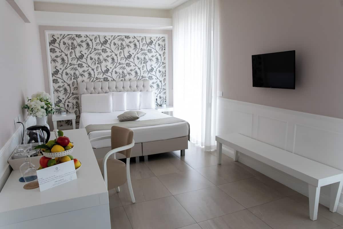 A modern hotel room with a bed, a desk with a fruit bowl, a chair, a bench, and a TV mounted on the wall.