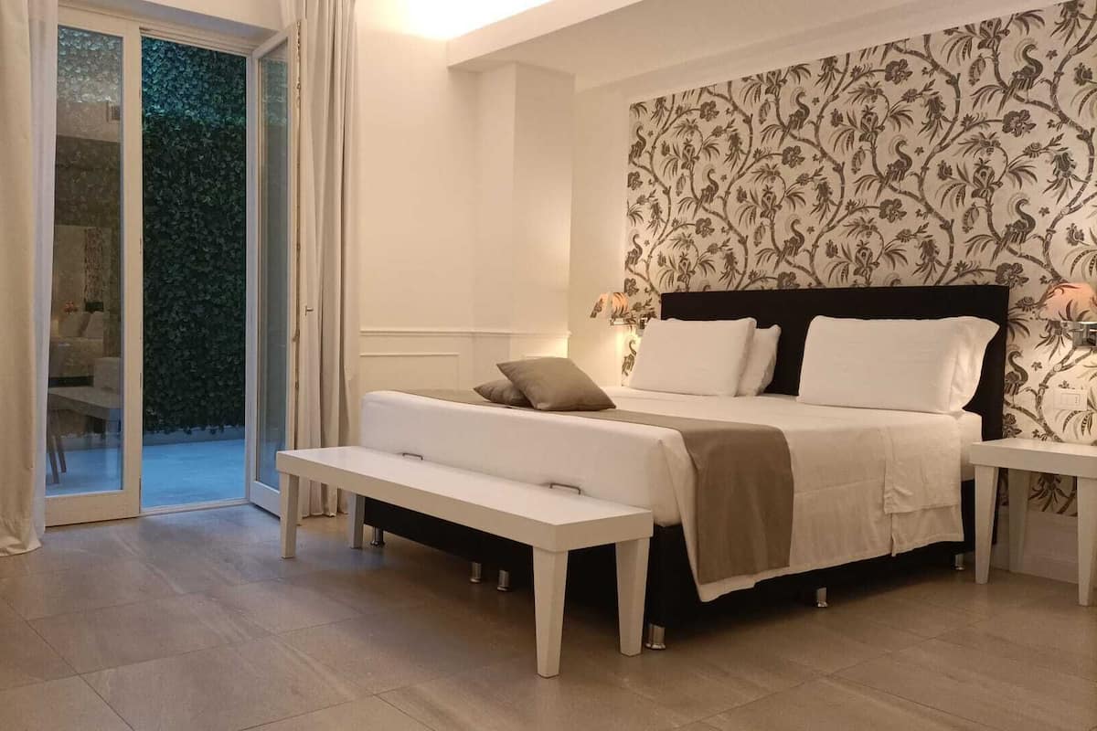 A bedroom with a bed, a bench, a nightstand, and a view of a garden through a glass door.