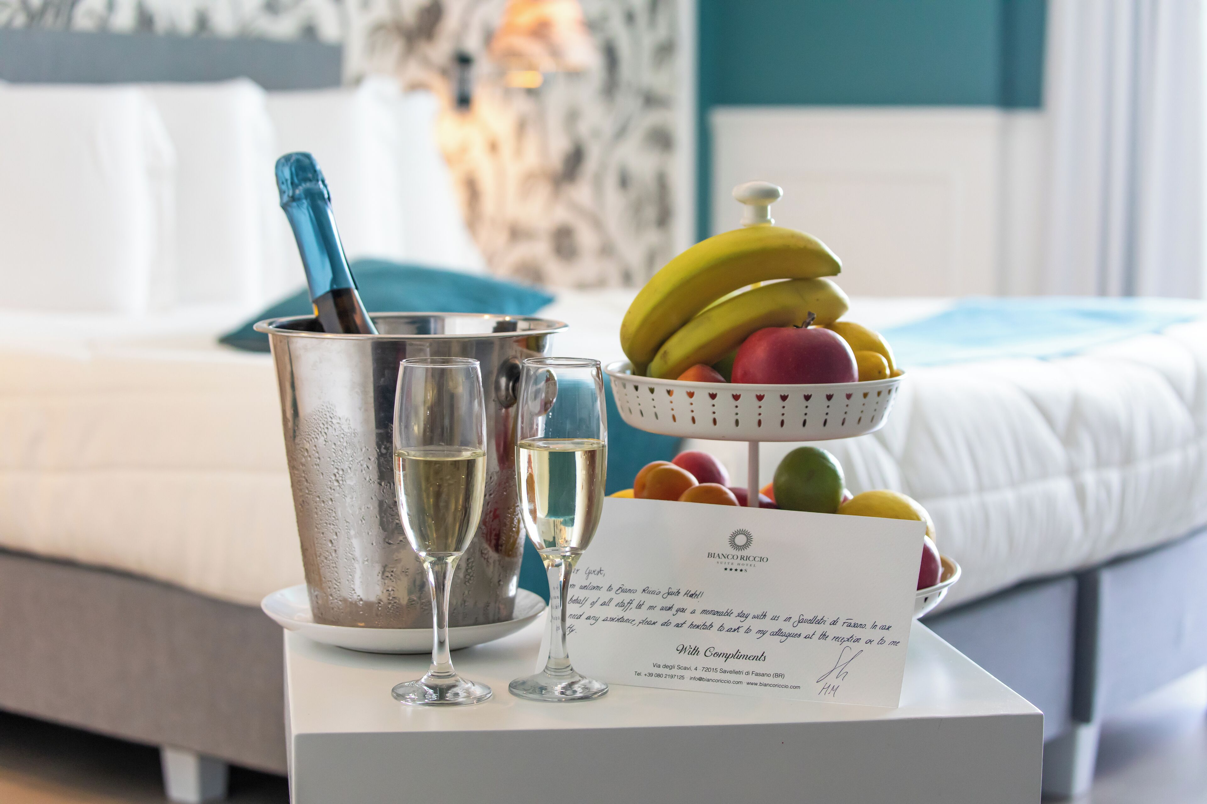 A hotel room with a bed, a bedside table with a champagne bottle, two glasses, and a fruit bowl.