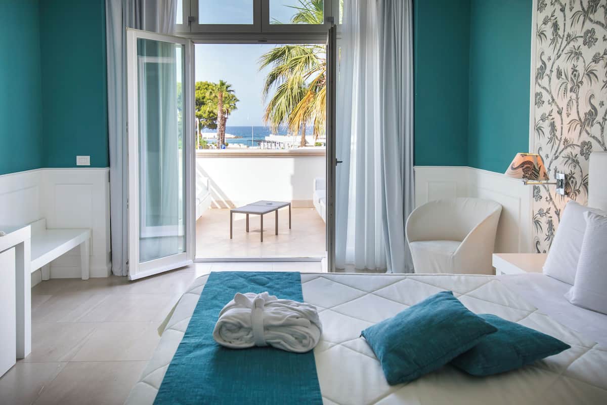 A hotel room with a bed, a bench, a table, and a view of the sea through a glass door.