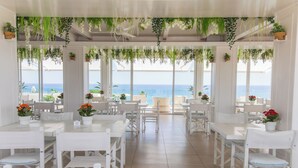 A restaurant with white tables and chairs, potted plants, and a view of the sea.