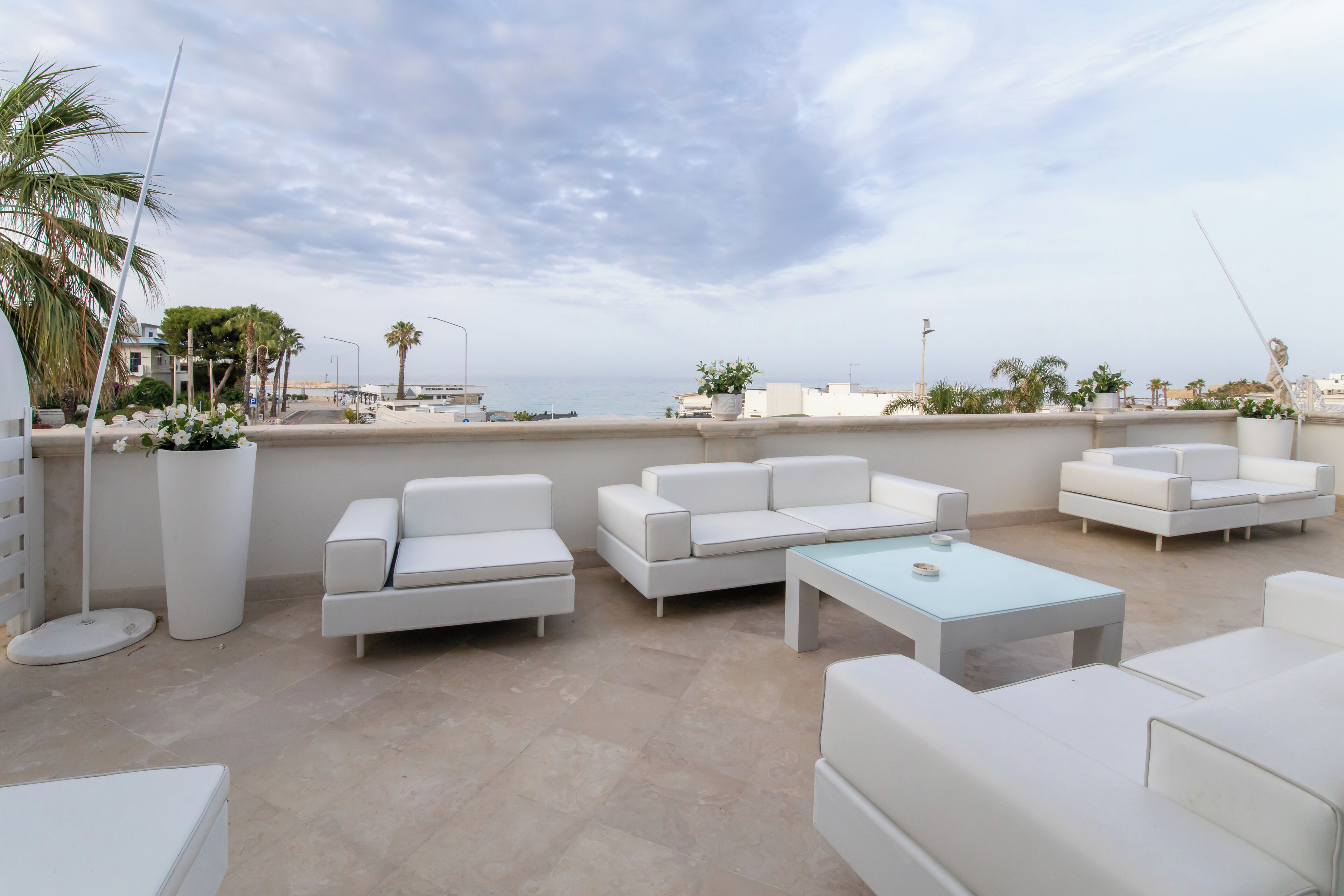 A rooftop terrace with white sofas and a light blue table, overlooking a coastal area with palm trees and buildings.