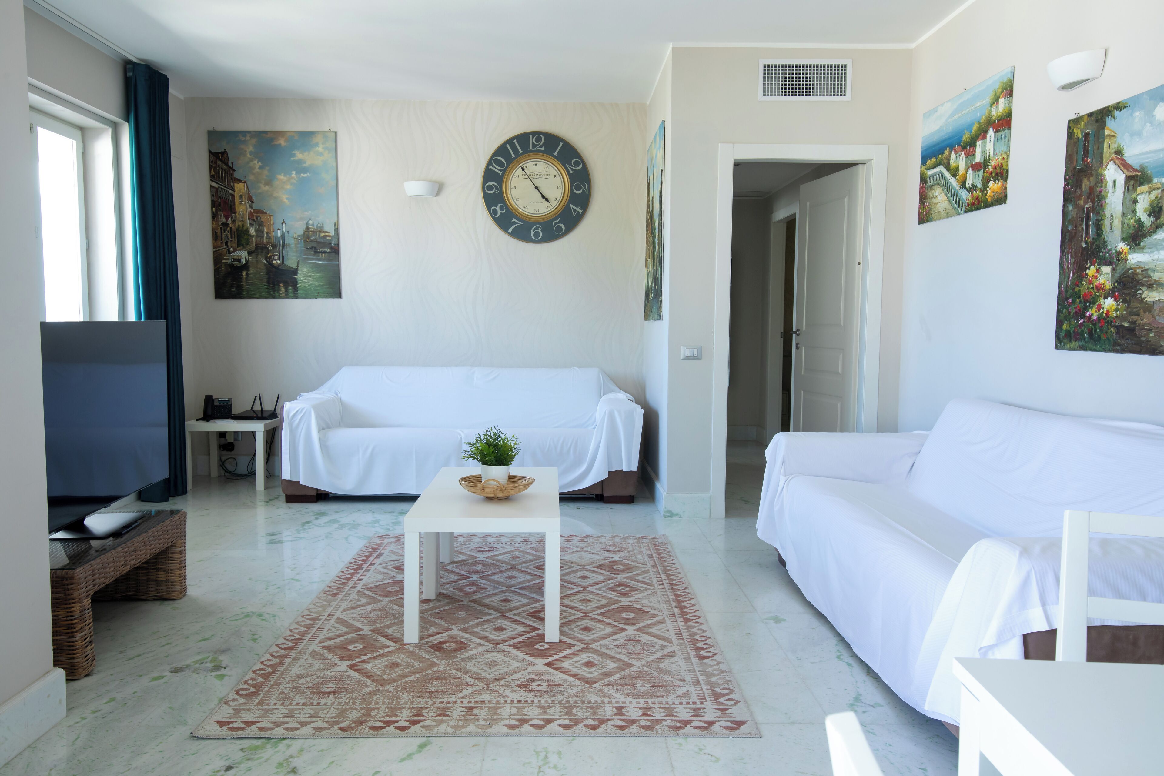 A living room with a white sofa, a coffee table with a plant, a clock on the wall, and artwork.