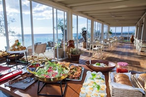 Una mesa de buffet con diversos platos, vistas al mar a través de amplios ventanales y un área de comedor informal con sillas y mesas.