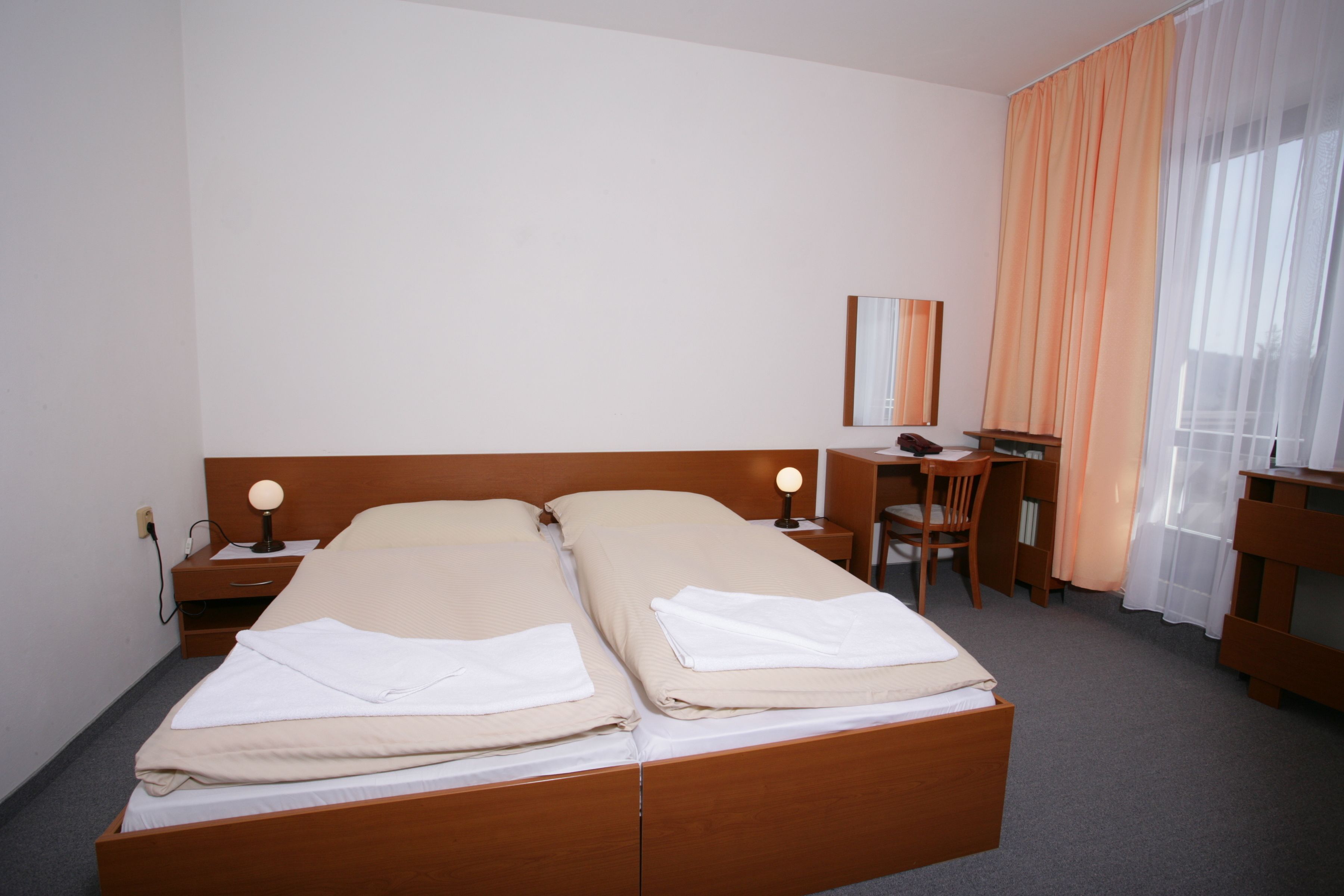 A double bed with two pillows, a bedside table with a lamp, a desk with a chair, and a window with curtains.