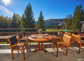 An outdoor seating area with a round wooden table and chairs, a vase with flowers, and a scenic view of trees and hills in the background.
