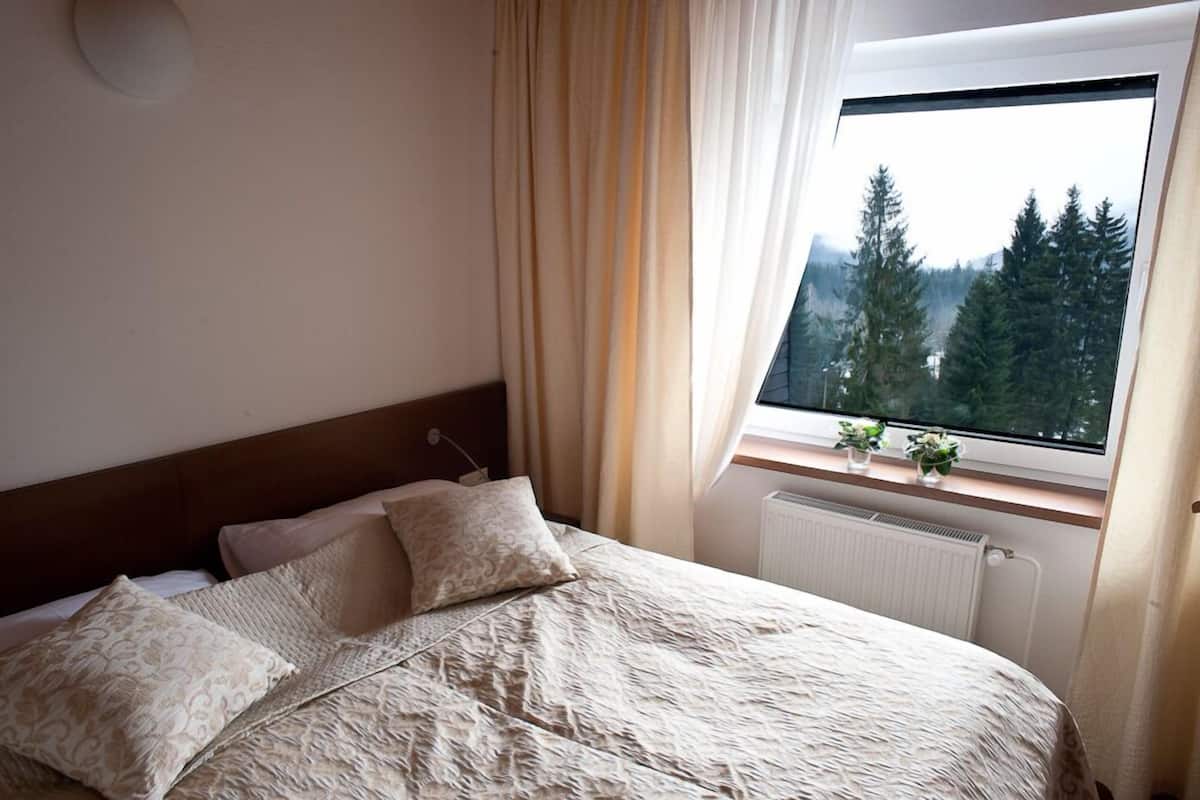 Un dormitorio con cabecera de madera, cama con ropa de cama estampada, ventana con vistas a árboles y un radiador debajo de la ventana.