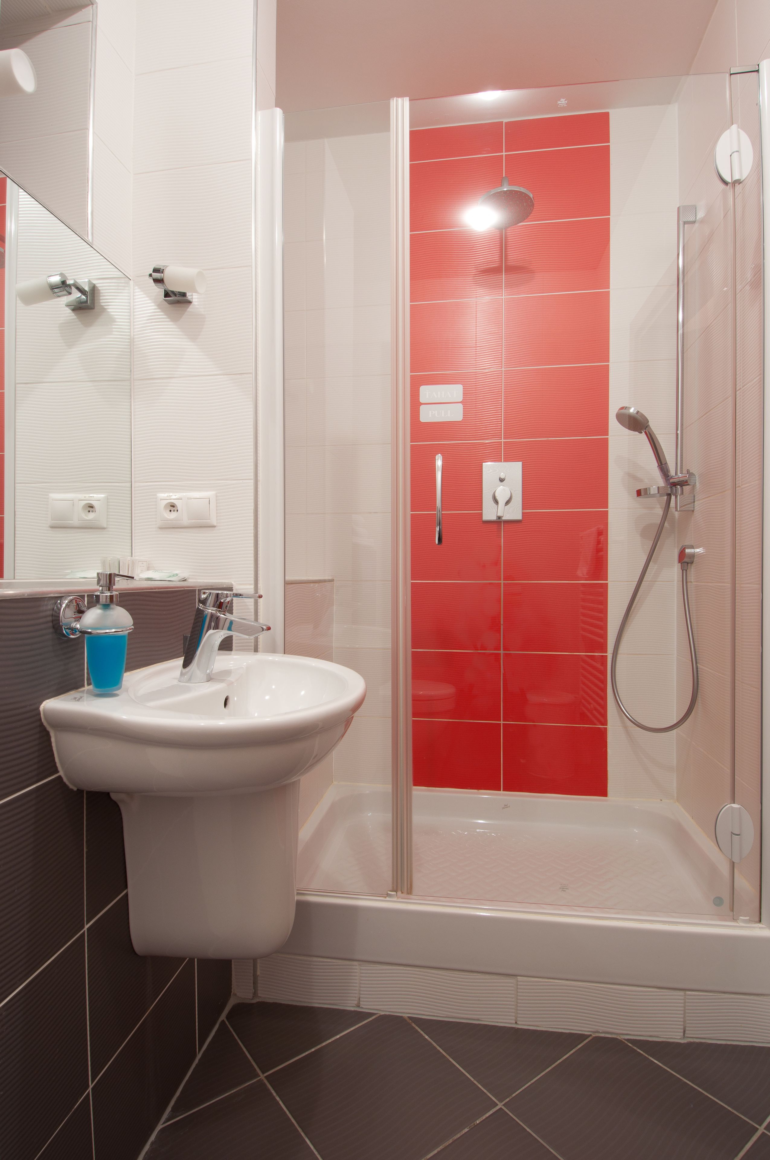 A modern bathroom with a white sink, a blue soap dispenser, a glass-enclosed shower, and a red-tiled wall.