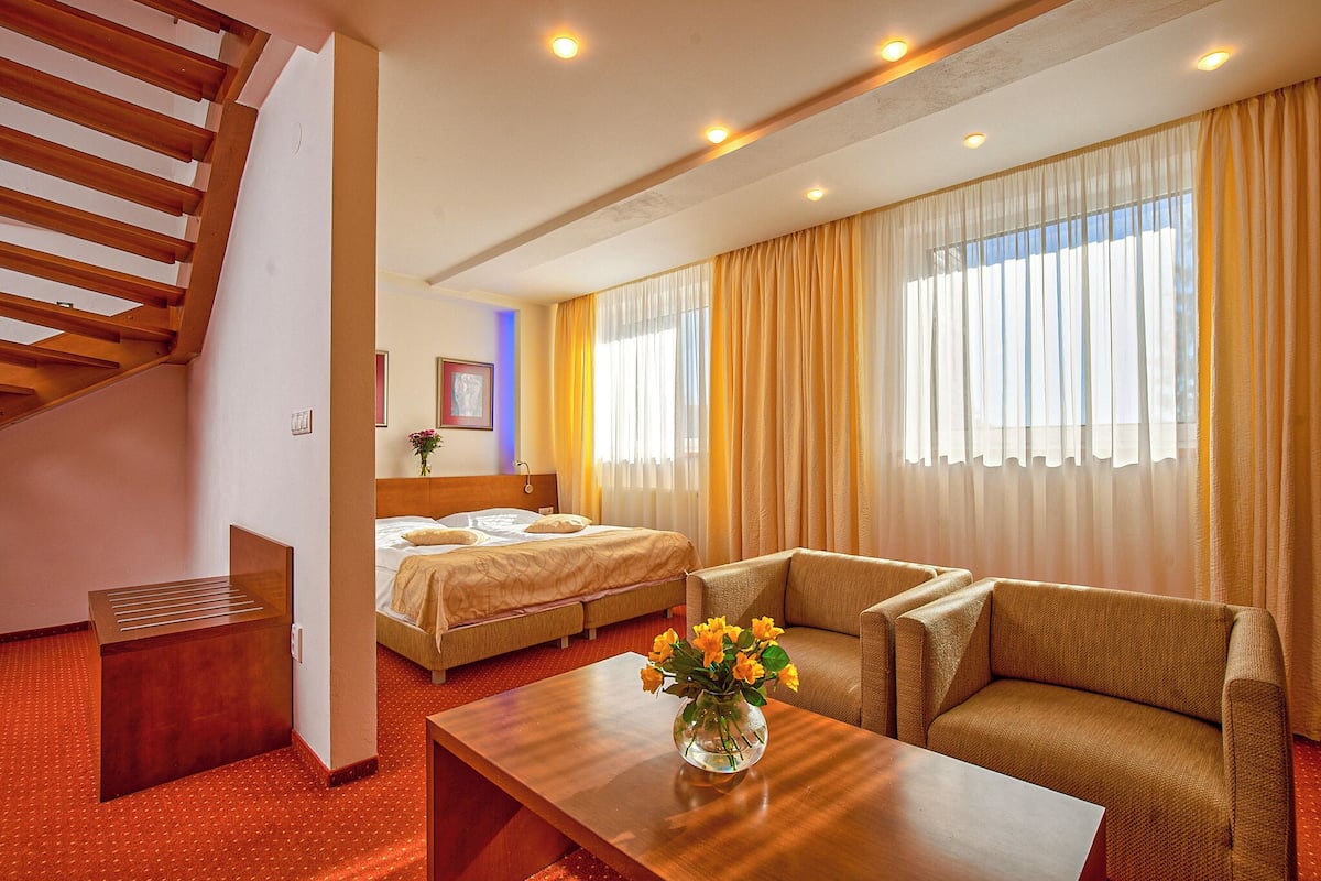 Habitación de hotel con escalera de madera, cama, sofá, mesa de centro con un jarrón de flores y ventanas grandes con cortinas.