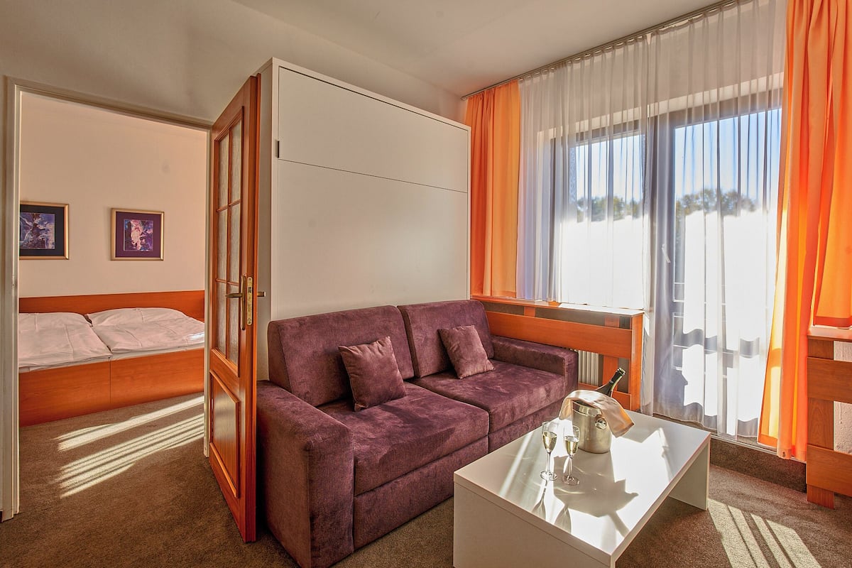 Una habitación de hotel compacta con un sofá púrpura, una mesita blanca con una botella y un vaso, una cama con sábanas blancas y una ventana con cortinas anaranjadas.