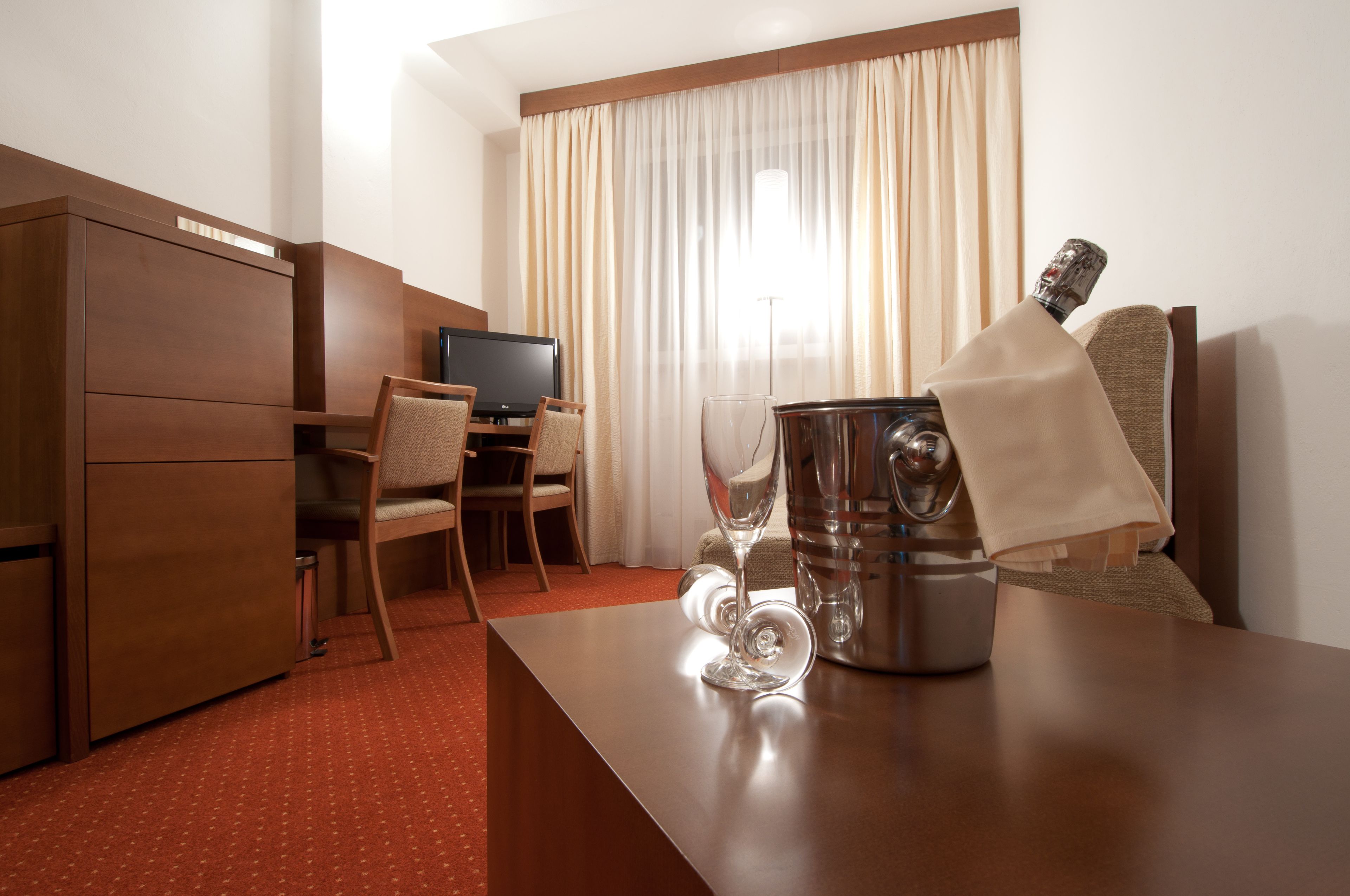 A hotel room with a wooden desk, a television, a minibar, and a large window with curtains.