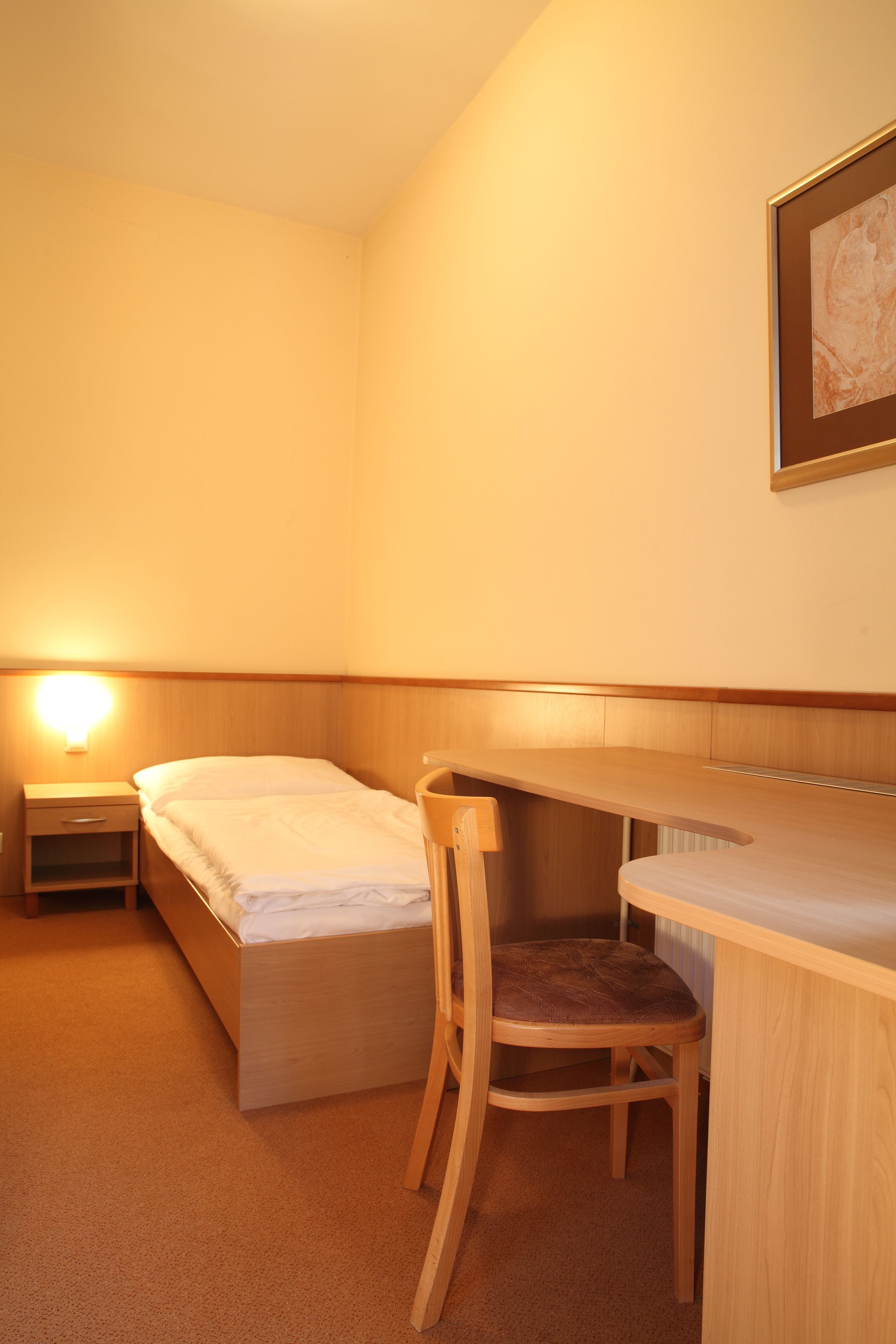 A small, well-lit room with a single bed, a desk, a chair, and a nightstand.