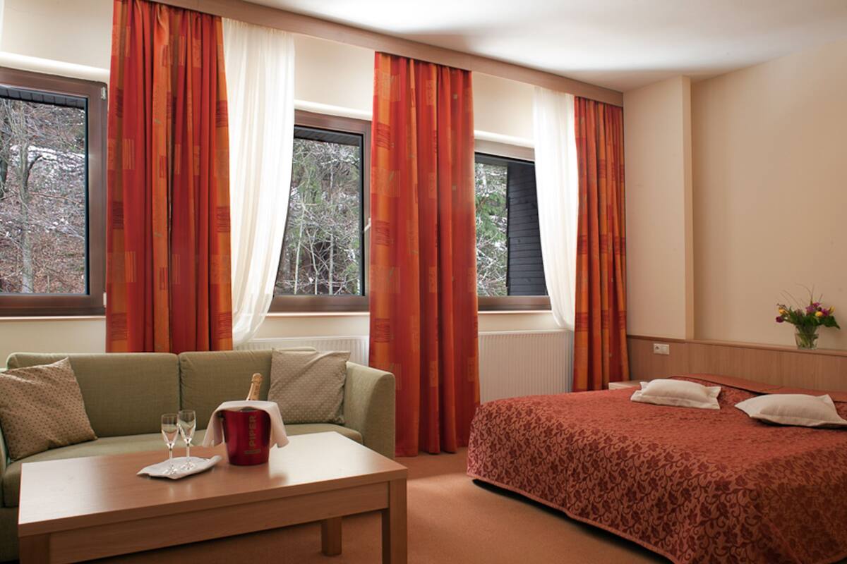 Una habitación de hotel con una cama, un sofá, una mesa con vasos y una botella, y cortinas.