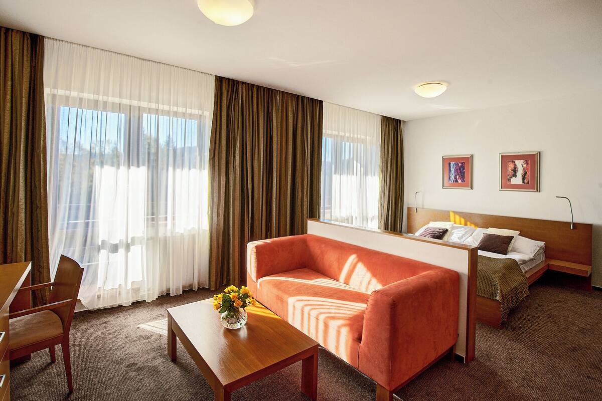 Habitación de hotel con sofá, cama, mesa de madera con un florero y un gran ventanal con cortinas.