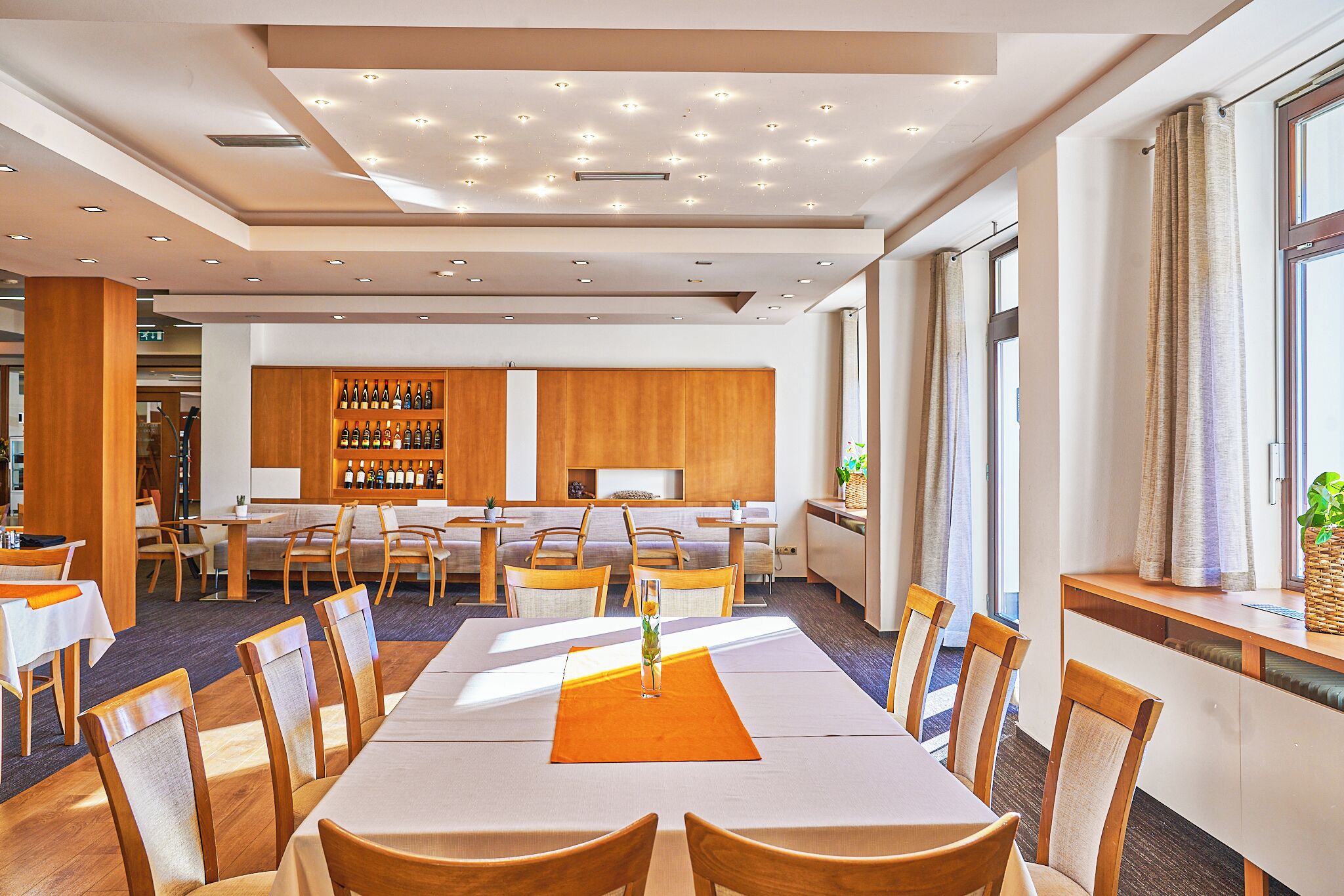 Uma sala de jantar de hotel com várias mesas e cadeiras, um bar com garrafas e uma janela grande com cortinas.