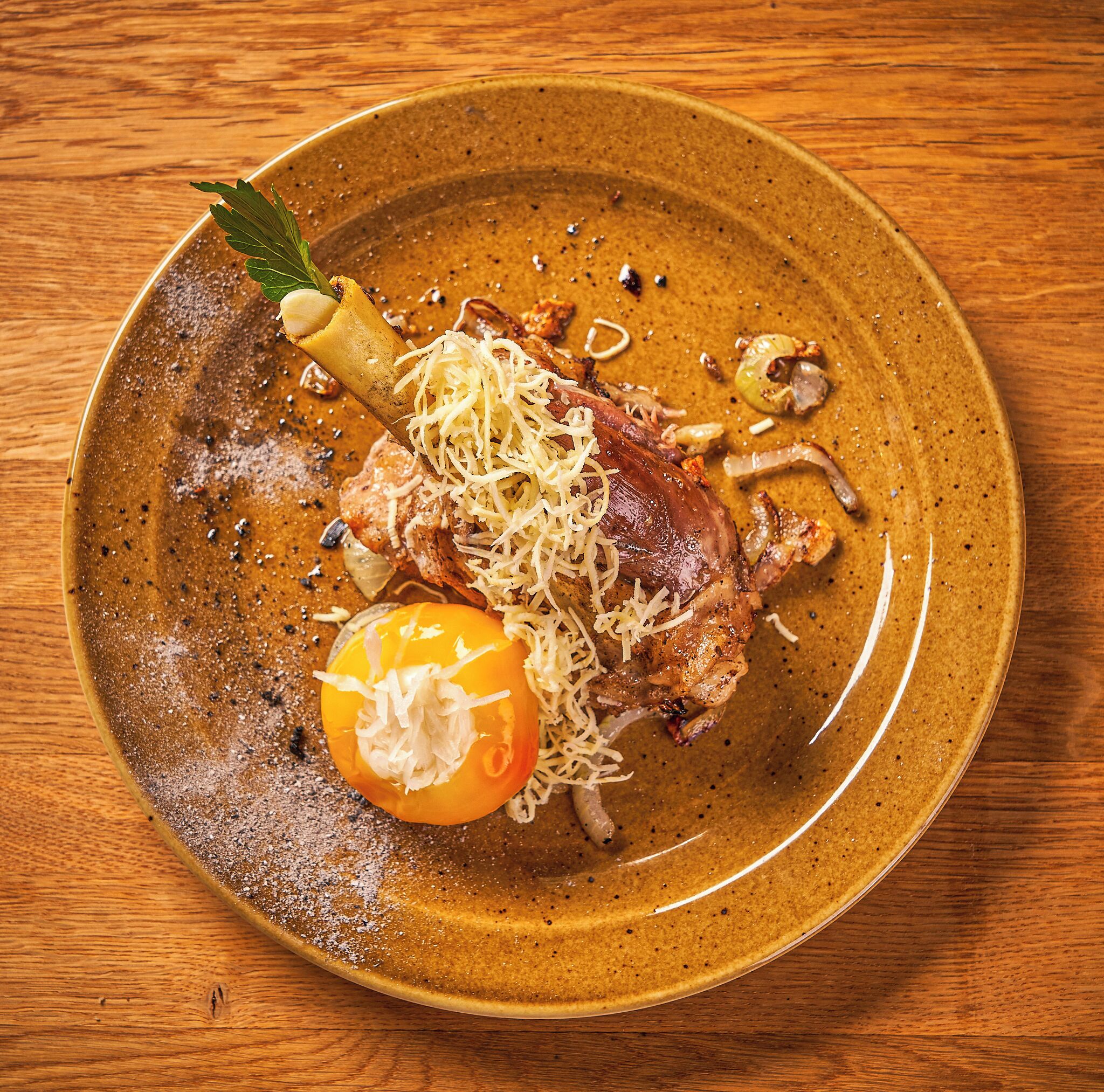 A plate with a piece of meat, a tomato, and some herbs on a wooden table.