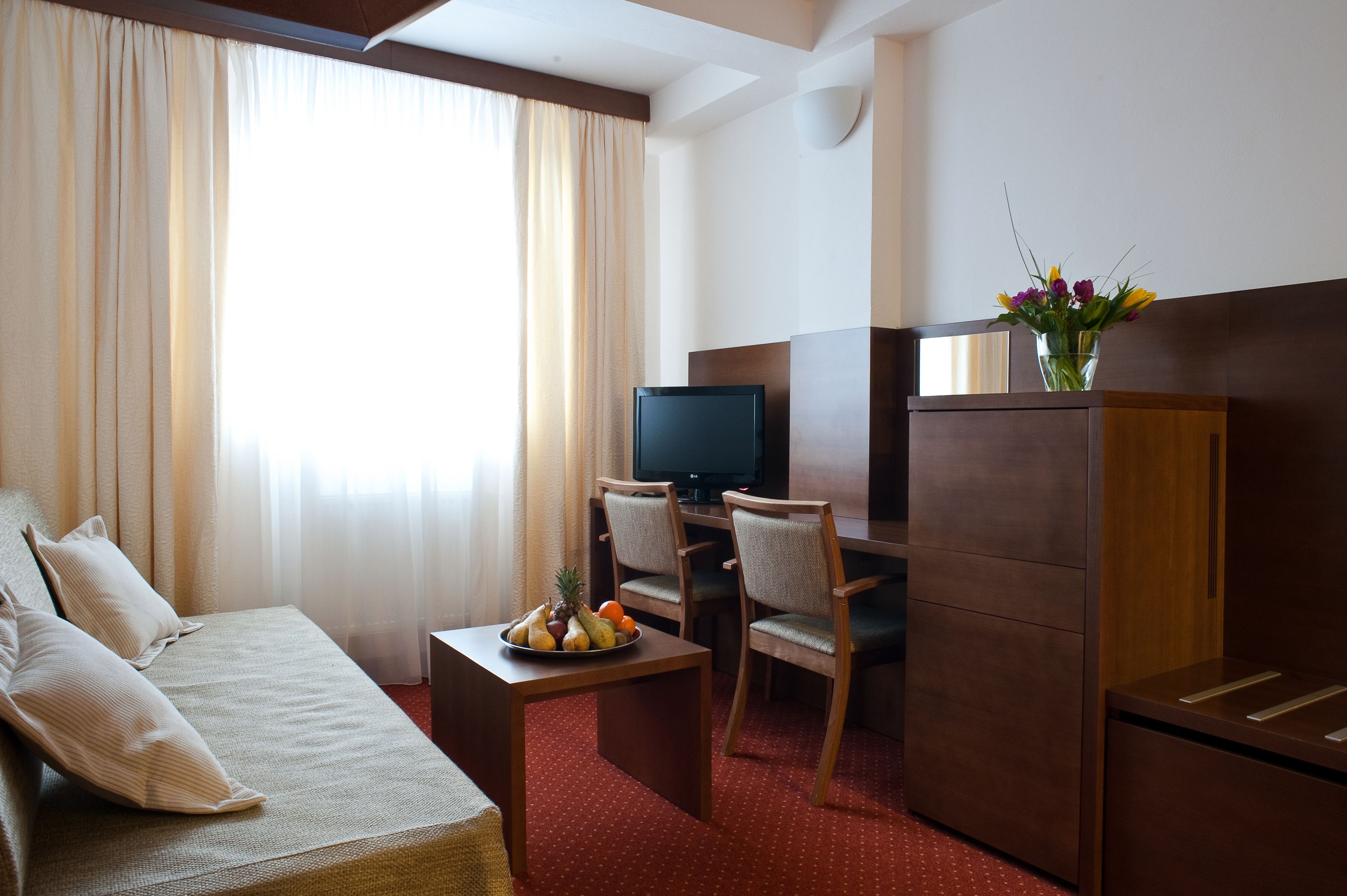 A hotel room with a bed, a desk with a computer, a chair, a small table with fruit, and a vase with flowers.
