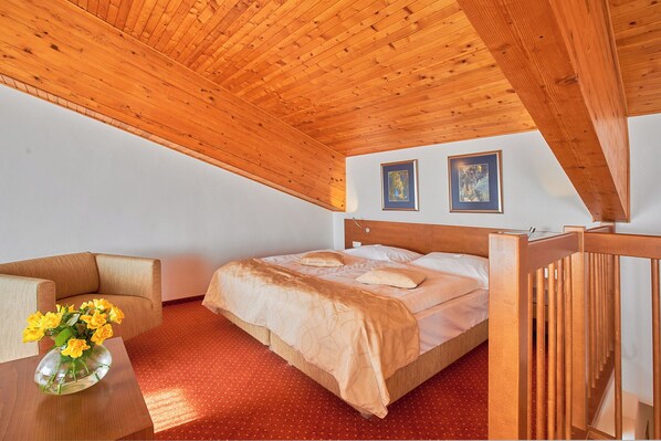 Un dormitorio con techo de madera, una cama con colcha color crema, una mesita de noche de madera, una silla y un jarrón con flores.