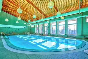 A large indoor swimming pool with a wooden ceiling, multiple windows, and sunlit areas.