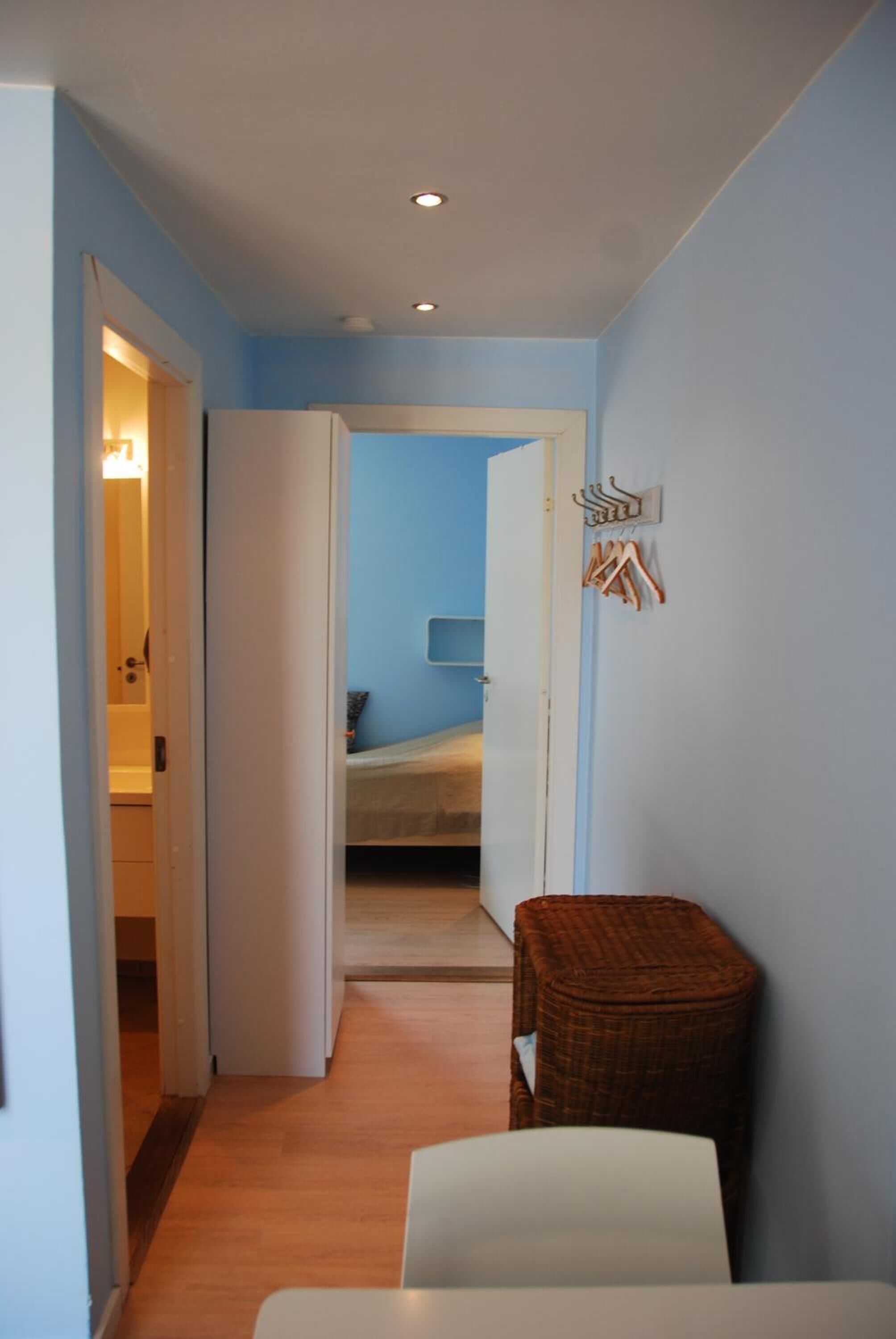 A narrow hallway with a bed in the background, a wicker chair, and a white table.