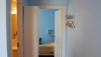 A narrow hallway with a bed in the background, a wicker chair, and a white table.
