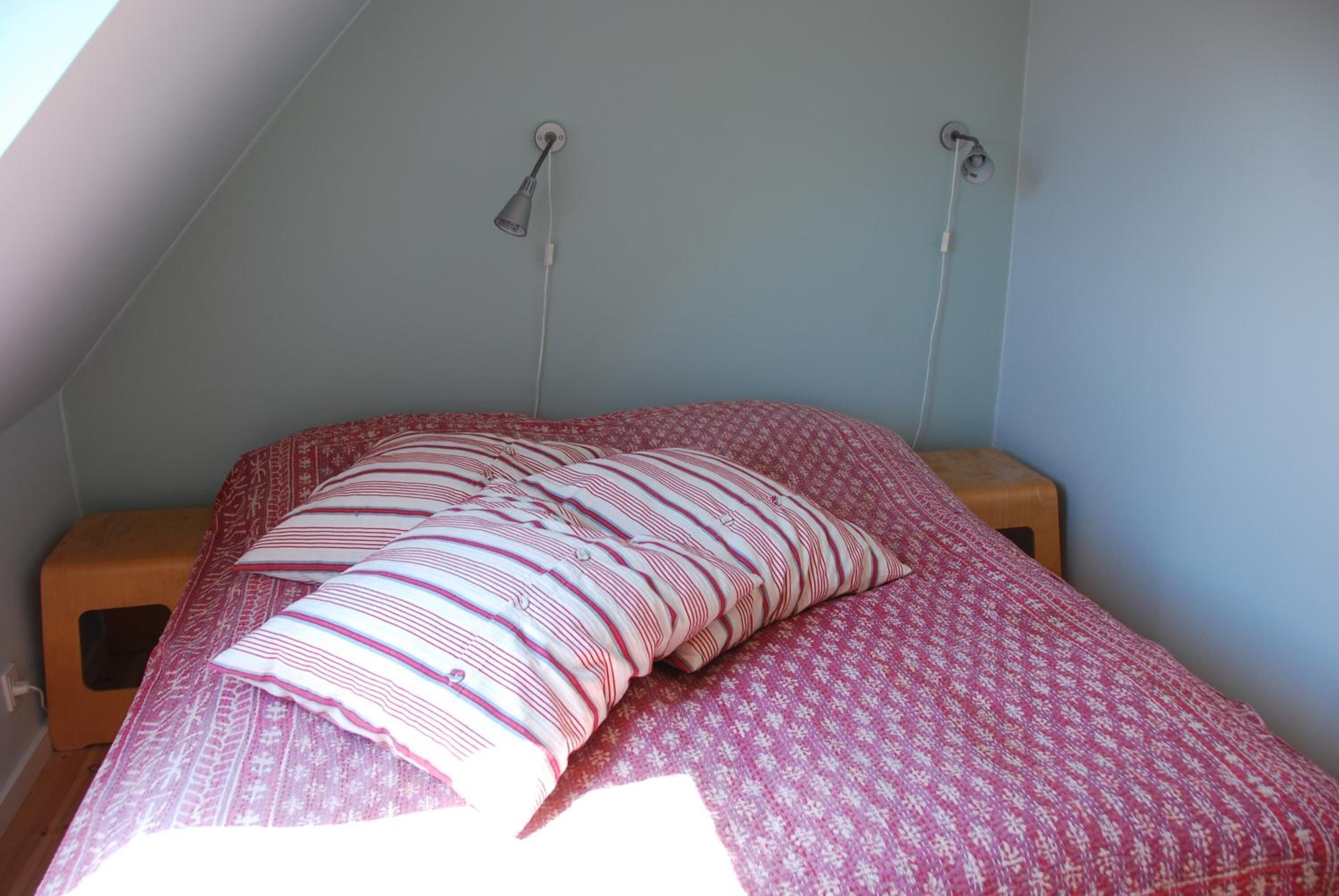 Un dormitorio con techo inclinado, una cama con colcha roja y blanca estampada, dos lámparas de noche y una mesita de noche de madera.