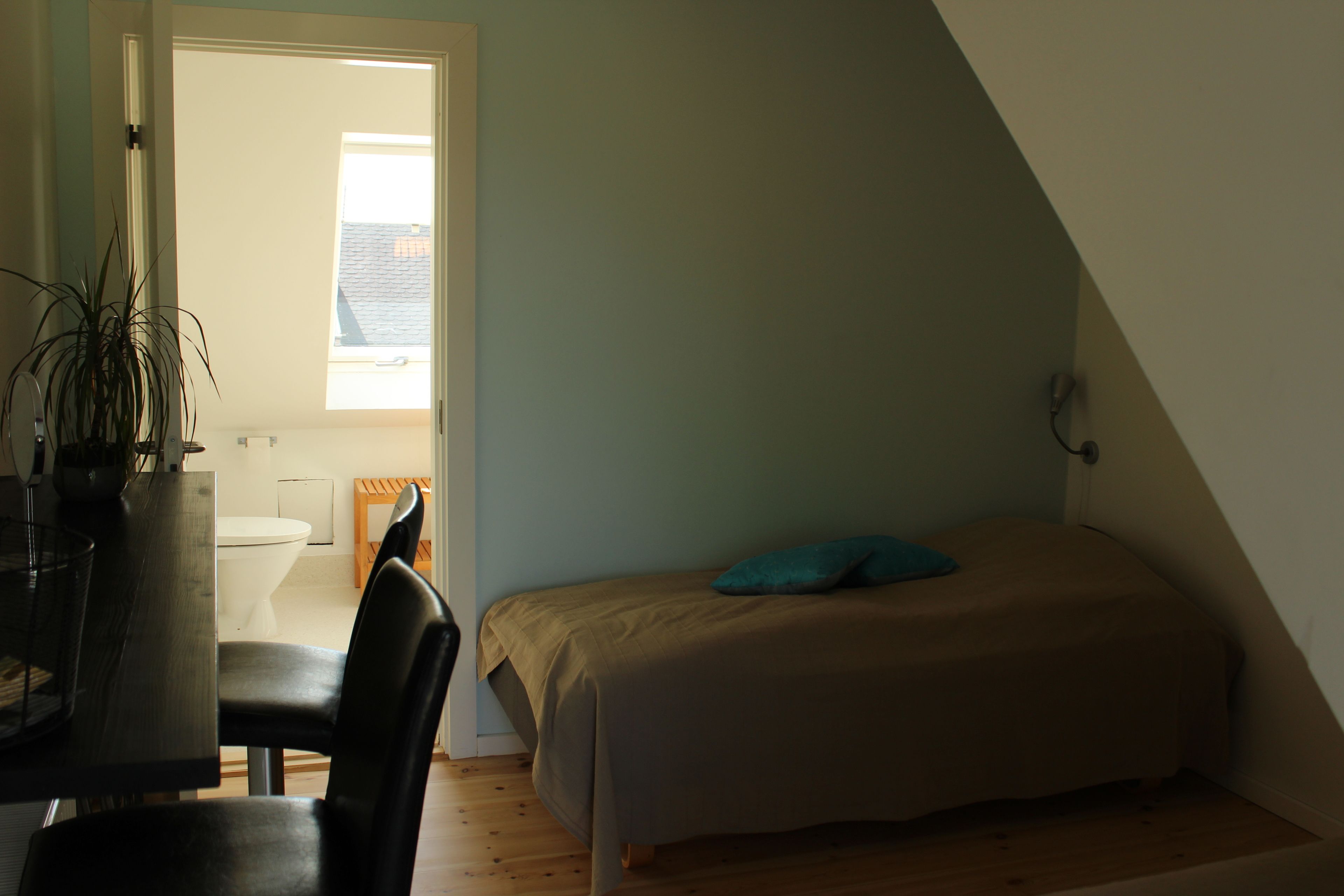 Un dormitorio pequeño y acogedor con una cama individual, suelo de madera, un escritorio con silla y ventana que proporciona luz natural.