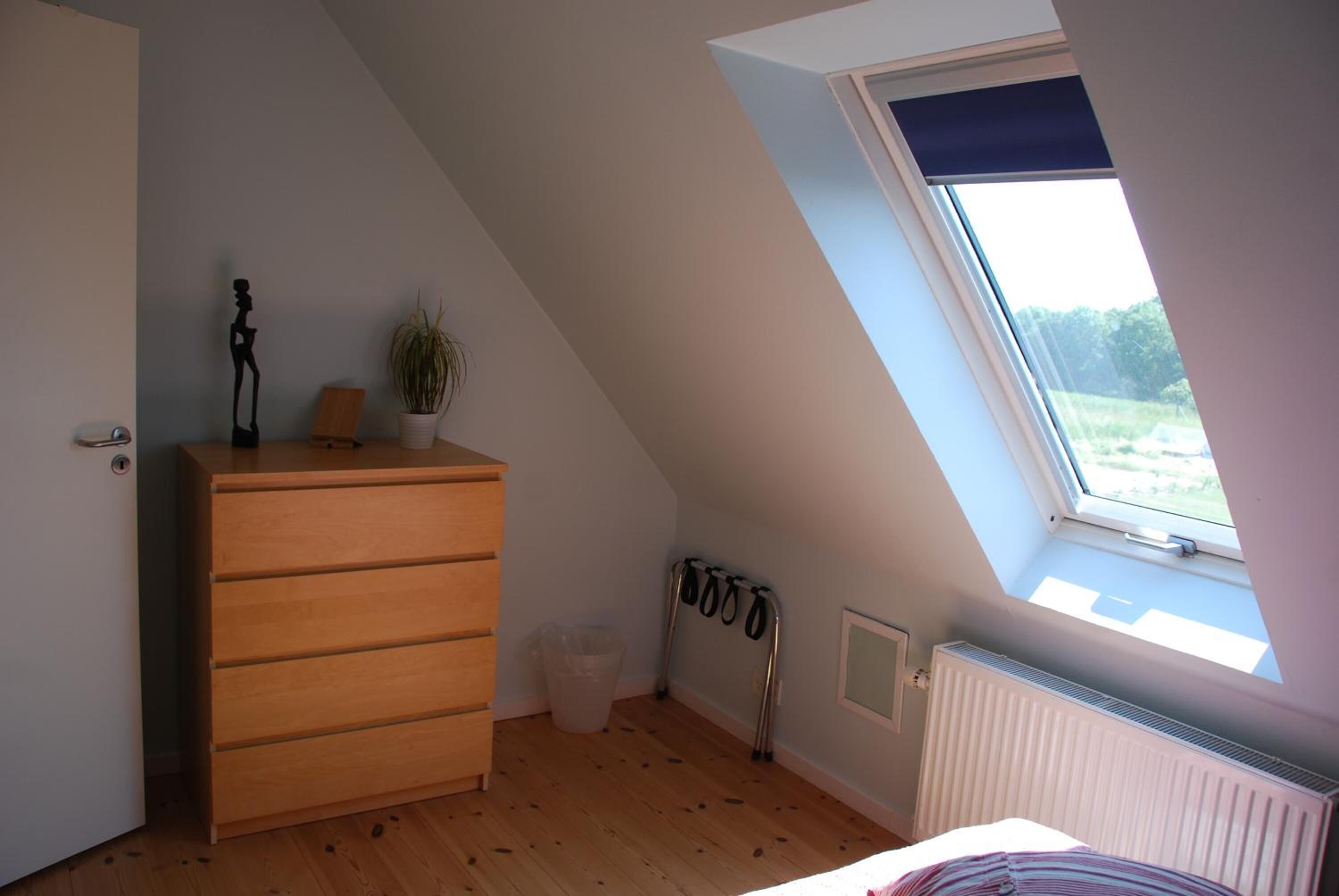 Et soveværelse med skråvægge, en trækommode, en radiator, et vindue med udsigt og et knagered.