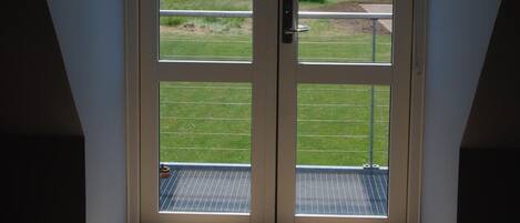 A room with a large sliding glass door leading to a balcony with a view of a wind turbine and green fields.