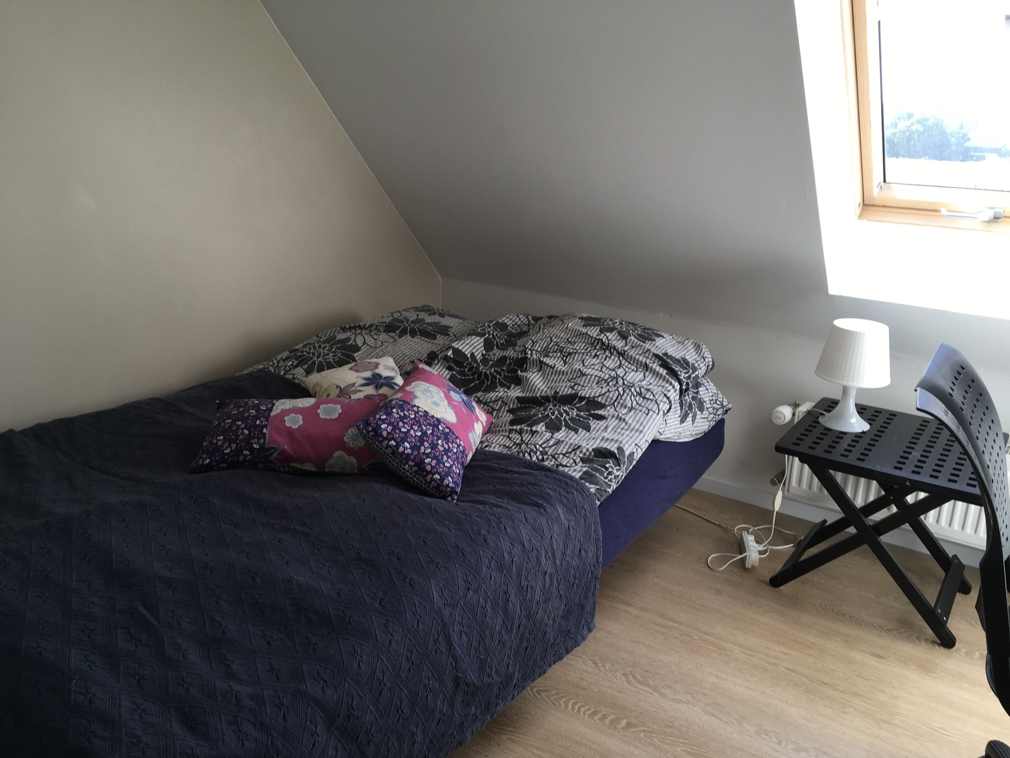 A bedroom with a bed, pillows, a desk with a lamp, and a window.