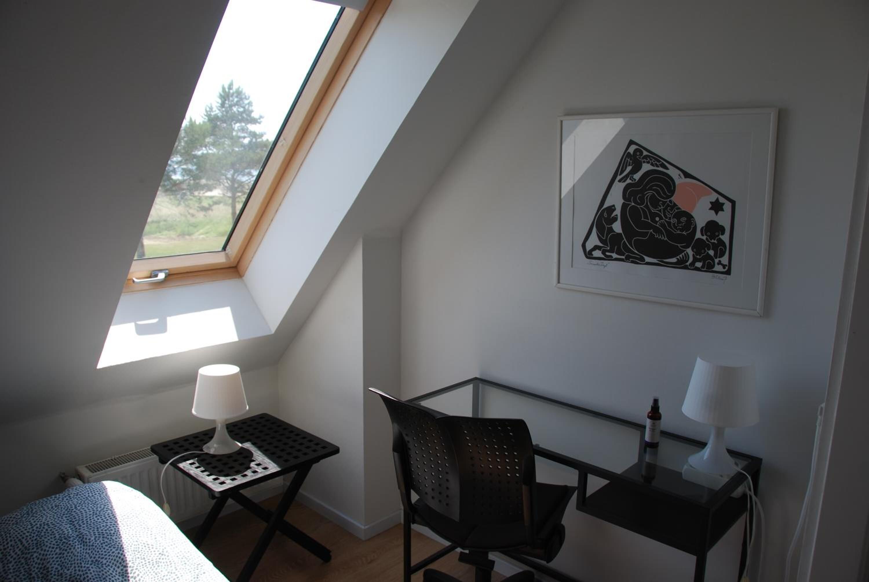 A bedroom with a skylight, a bed, a desk with a lamp, a chair, and a framed picture on the wall.