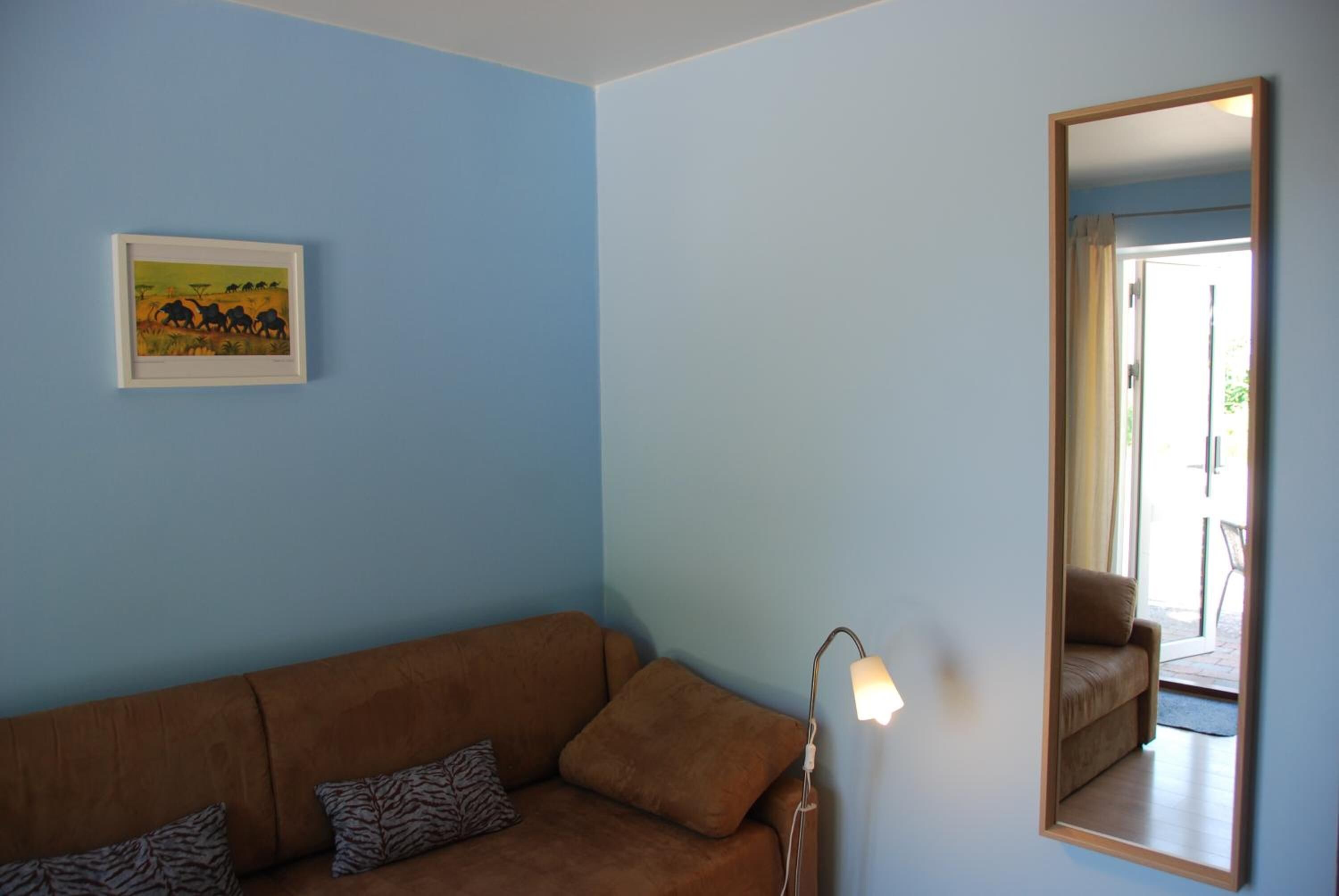 A room with a brown sofa, a framed picture on the wall, a standing lamp, and a mirror reflecting a door and a chair.