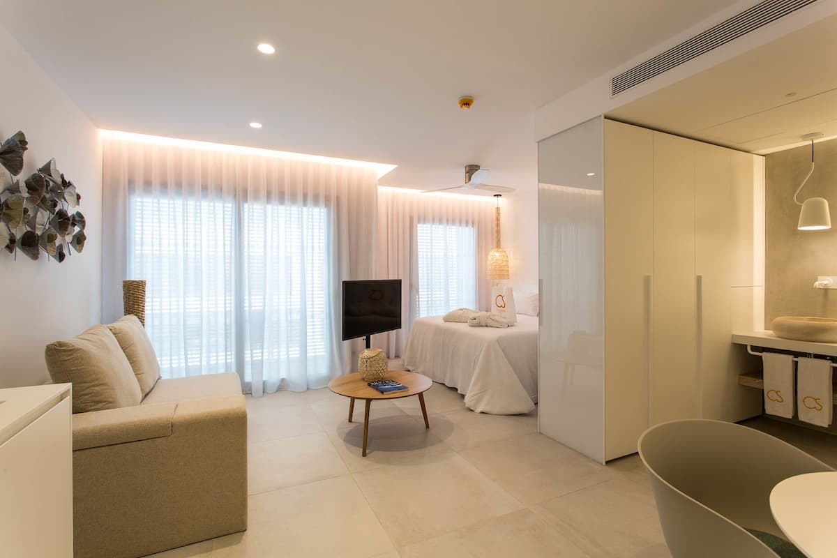 A modern living room with a sofa, a round coffee table, a flat-screen TV, and a bed with a white duvet.