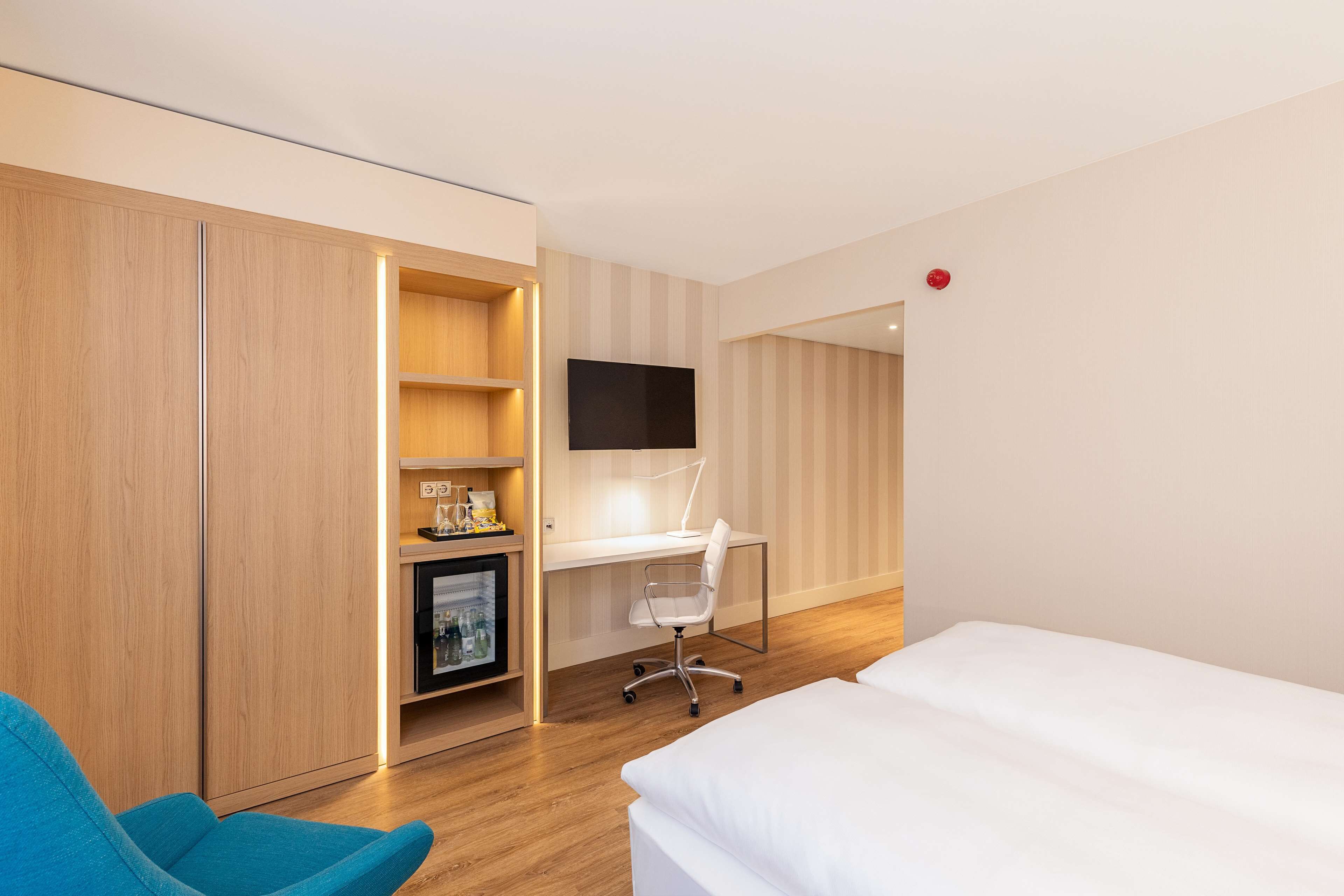 A hotel room with a bed, a desk with a chair, a TV, and a minibar.