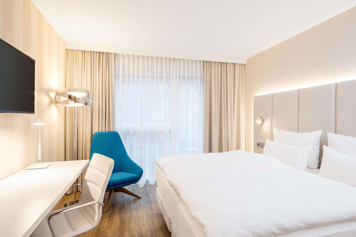 A hotel room with a bed, a desk, a chair, a TV, and a window with curtains.