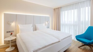 A modern hotel room with a large bed, a small side table with a vase, a blue chair, and a large window with sheer curtains. - NH Mannheim (Mannheim)