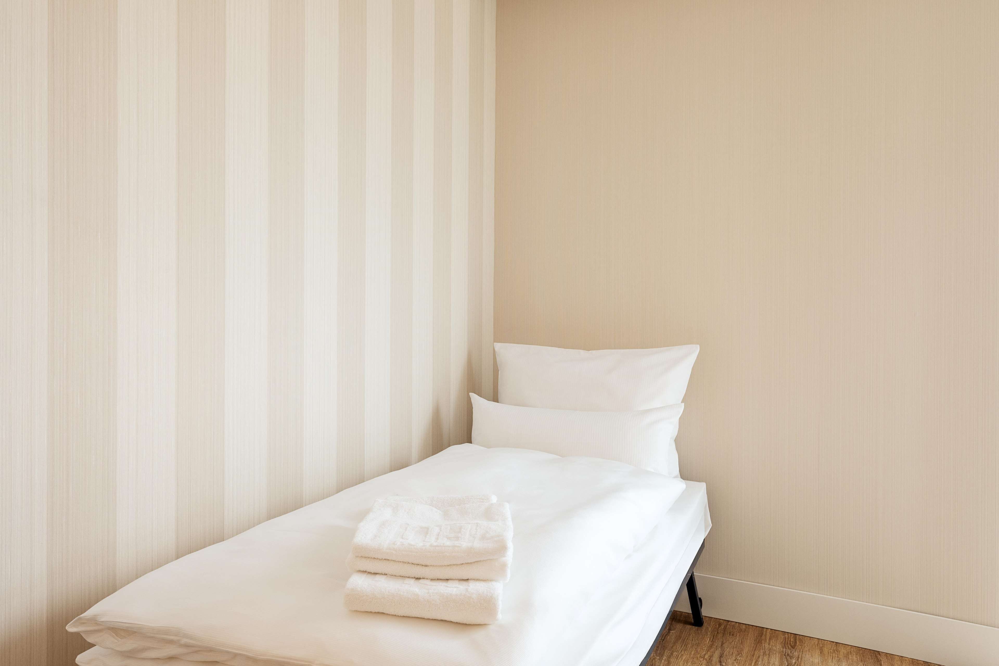A neatly made bed with white linens and pillows, a striped curtain, and a wooden floor.