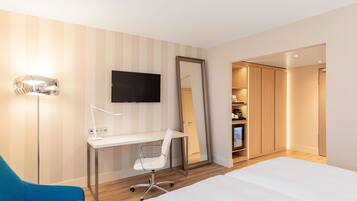 Habitación de hotel con una cama, un escritorio con silla, televisor, un espejo grande y un armario.