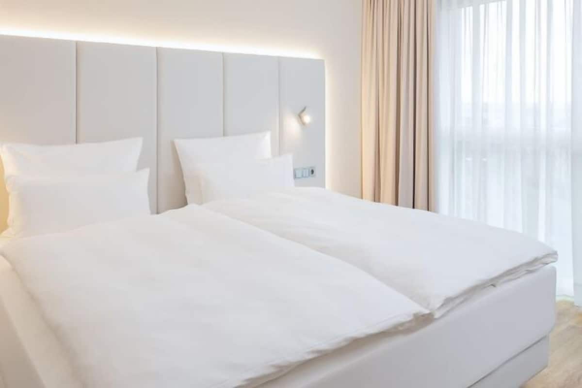 A modern hotel room with a large bed, a headboard with integrated lighting, and a view through sheer curtains.