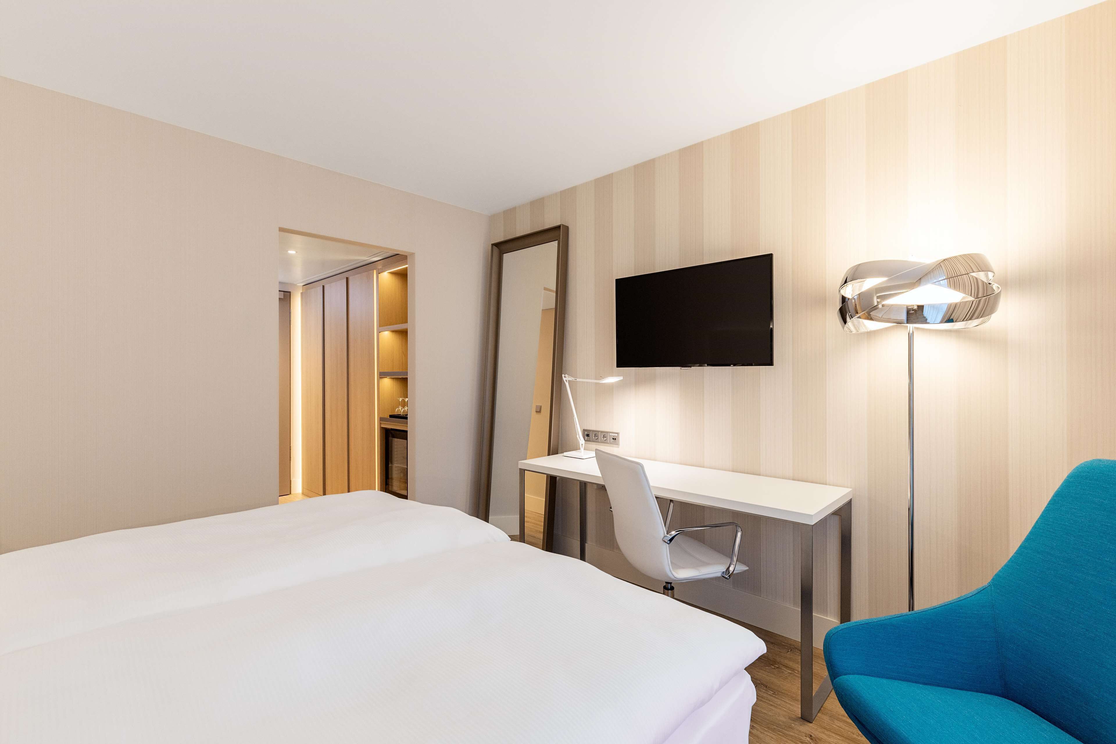 A hotel room with a bed, a desk, a chair, a TV, and a floor lamp.