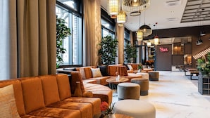 A modern lounge area with sofas and ottomans, large windows, and hanging pendant lights. - NH Mannheim (Mannheim)