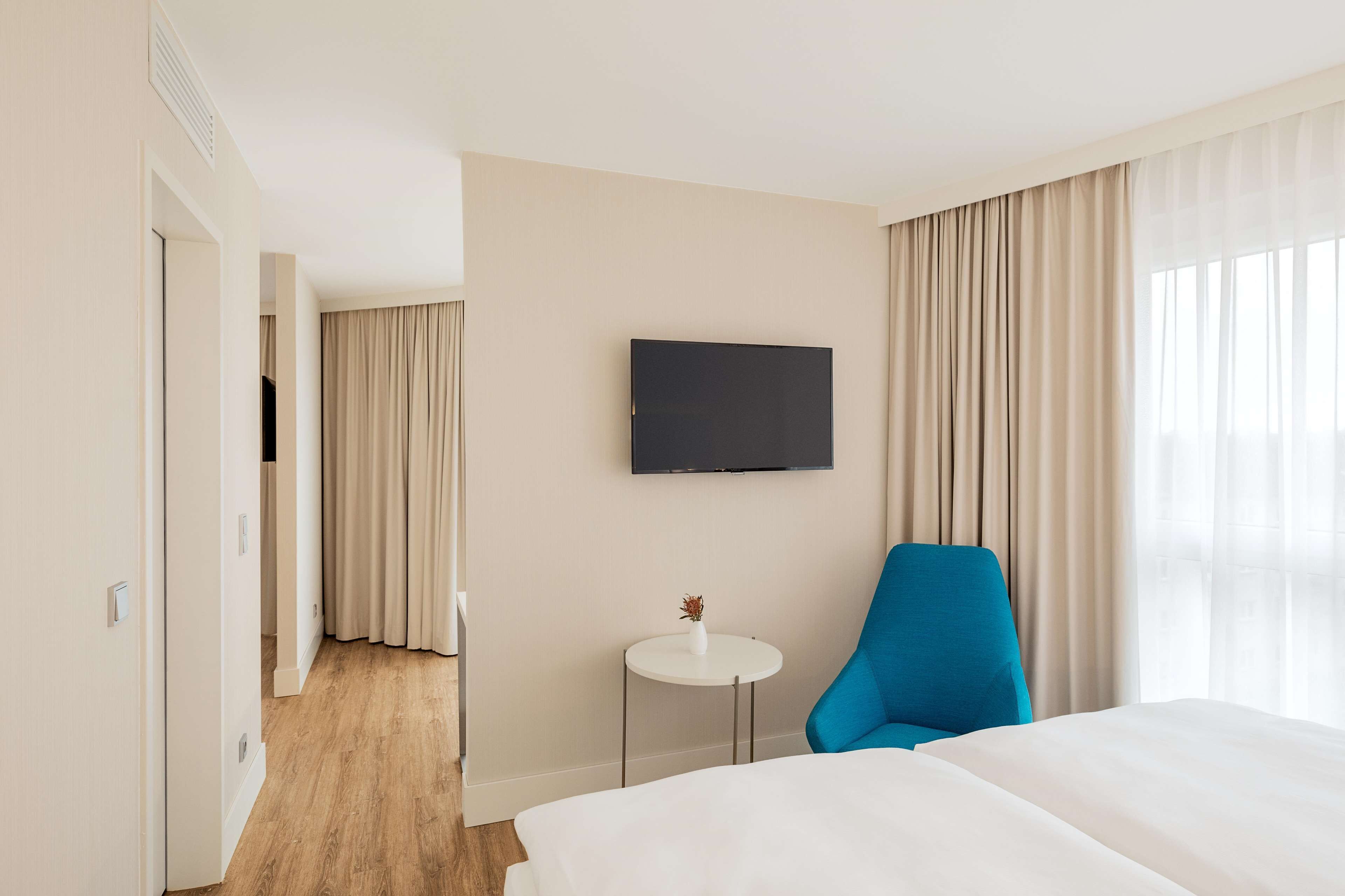 Une chambre d’hôtel avec un lit, un fauteuil bleu, une petite table avec un vase et un téléviseur fixé au mur.