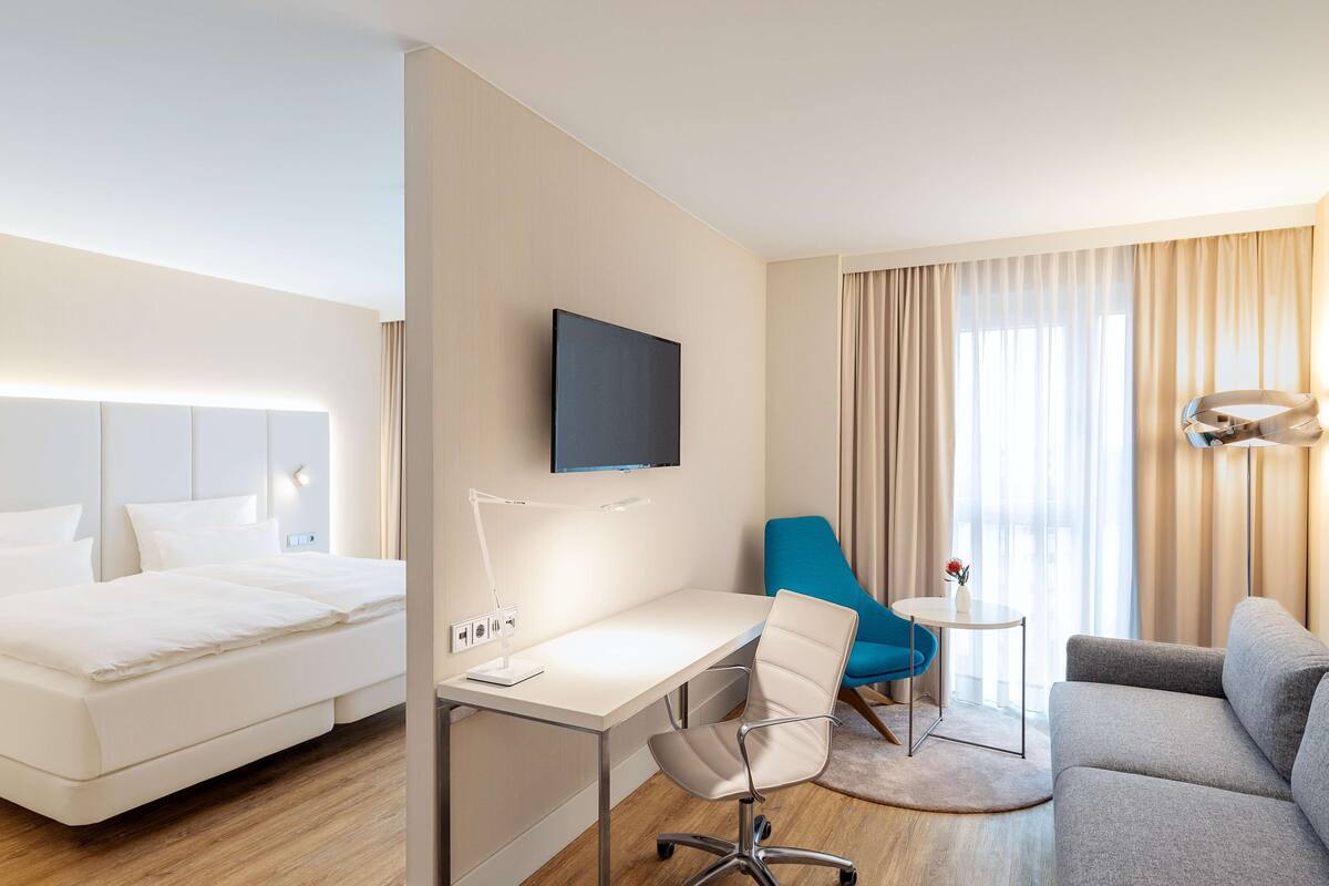 A hotel room with a bed, desk, chair, sofa, TV, and curtains.