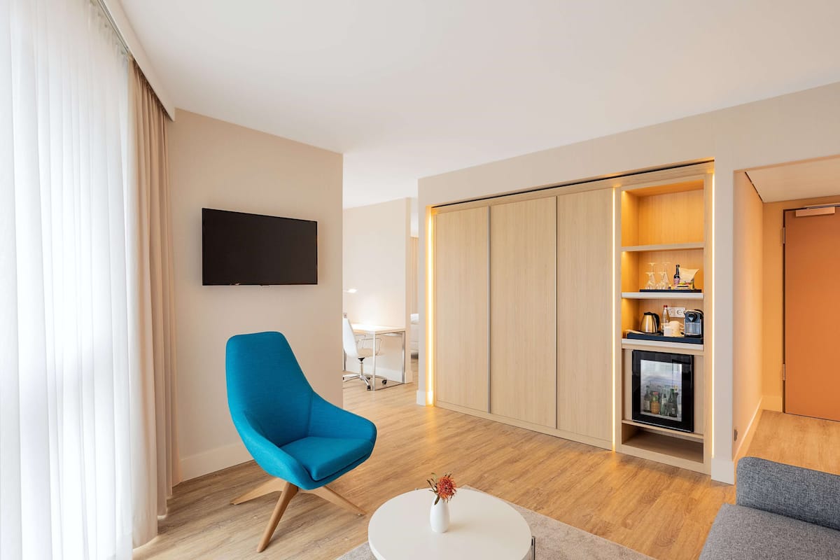 A modern hotel room with a blue armchair, a white coffee table with a vase, a flat-screen TV, a sliding wardrobe, and a minibar.