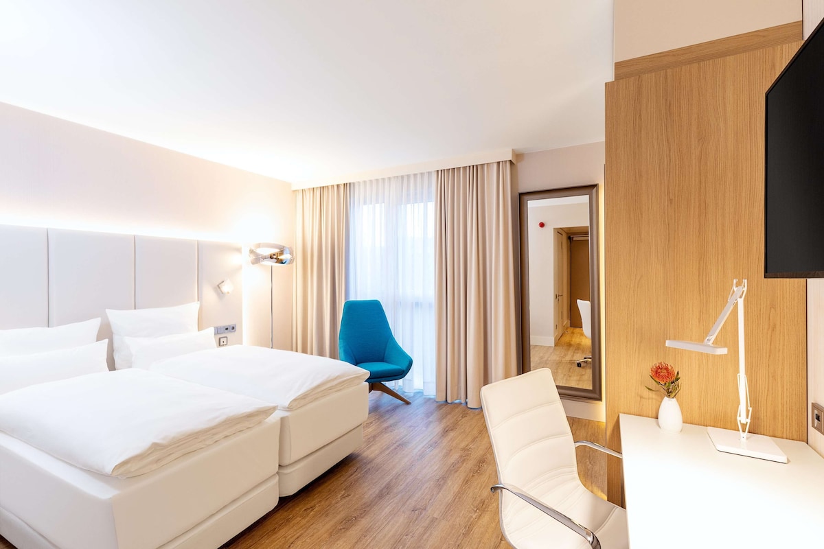 A hotel room with a bed, a desk, a chair, a blue armchair, and a mirror.
