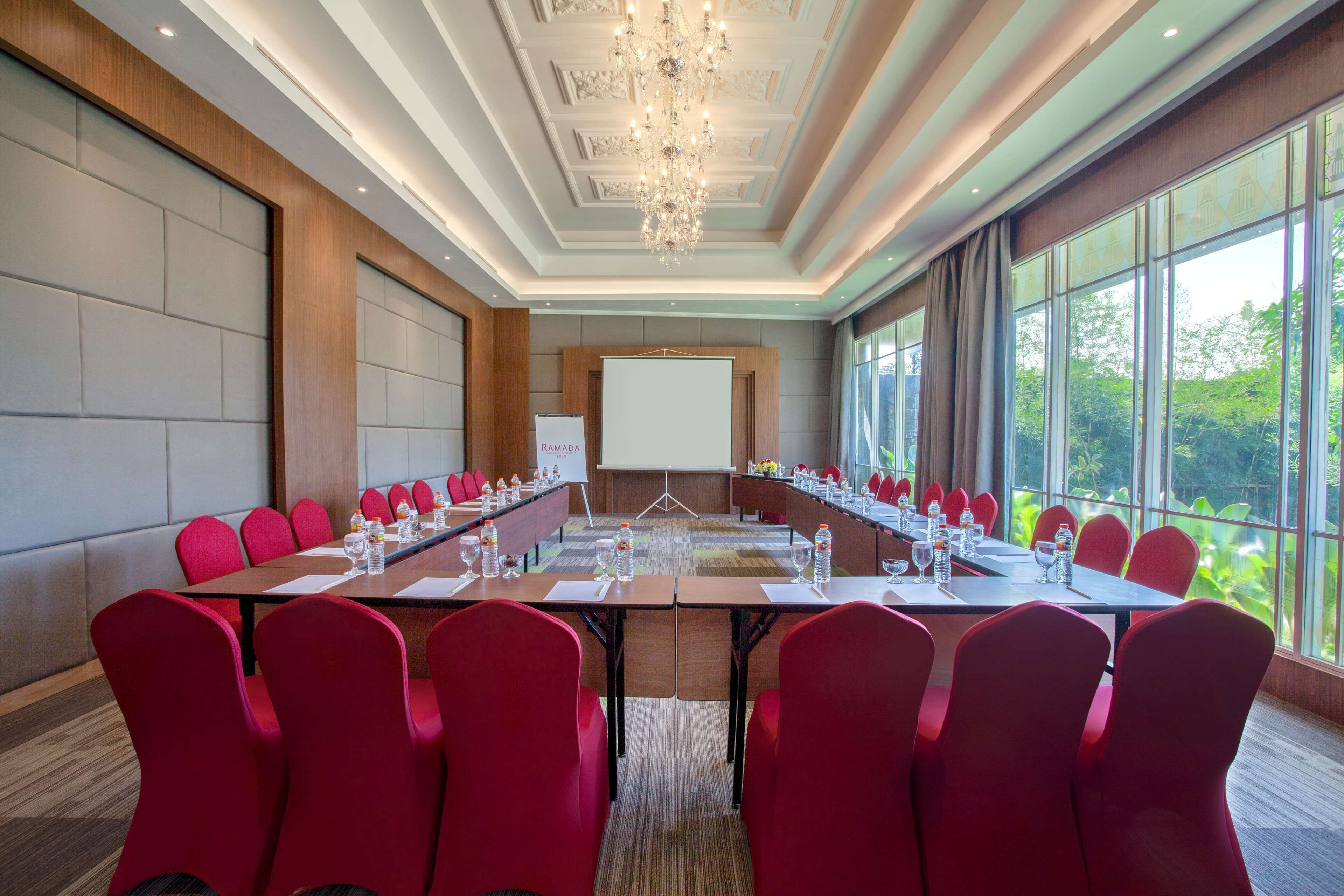 Un salón de conferencias con una larga mesa, sillas rojas, una pantalla de proyector y ventanas amplias con vistas a zonas verdes.