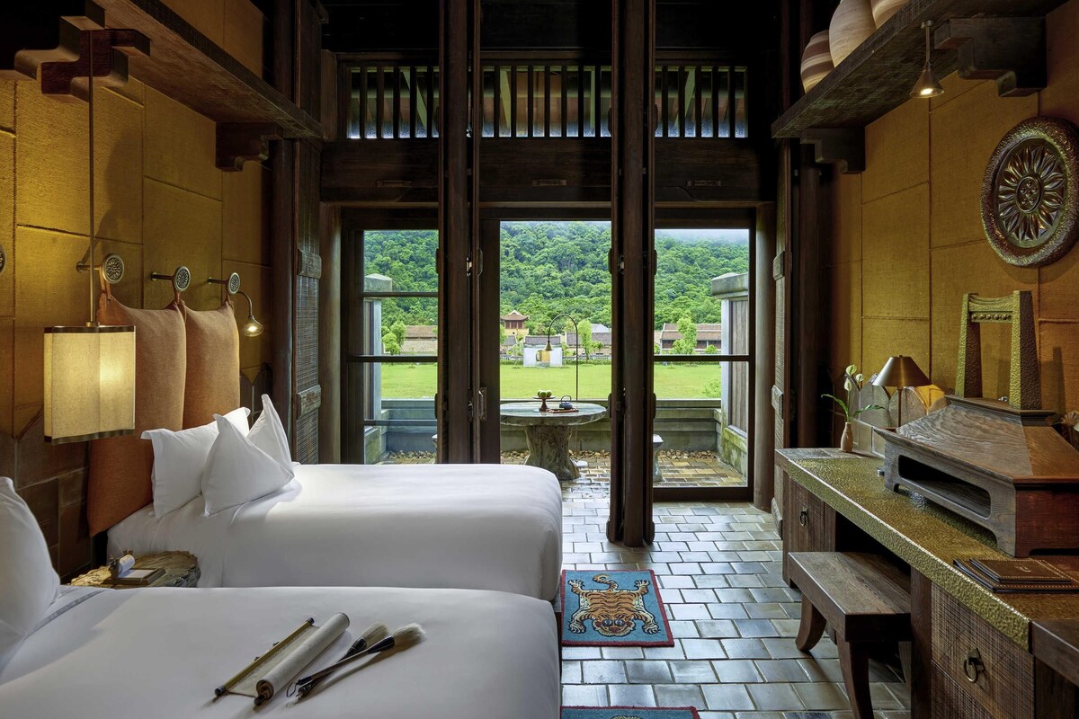 A hotel room with two beds, a desk, a TV, and a view of a green landscape.
